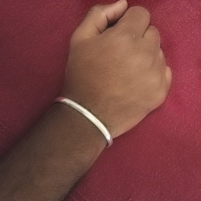 May include: A smooth, polished silver-toned cuff bracelet with a rounded profile. The bracelet features a continuous, solid band design that encircles the wrist, showcasing a reflective metallic surface that catches the light evenly across its curved exterior.