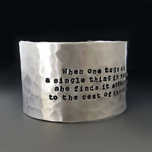 May include: A wide, hammered silver cuff bracelet. The bracelet has a textured surface and features the quote: "When One tugs at a single thing in nature, she finds it attached to the rest of the world."