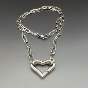 May include: A silver-toned necklace featuring a chain of elongated oval links and a prominent heart-shaped pendant. The pendant is constructed from a thick, metallic frame that mimics the appearance of a carabiner clasp, complete with a spring-loaded gate.