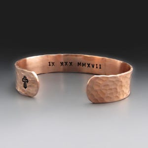 May include: A copper cuff bracelet with a hammered texture. The interior features the black engraved Roman numerals IX XXX MMXVII and a small black cross symbol on the left side. The bracelet has an open design with rounded ends.