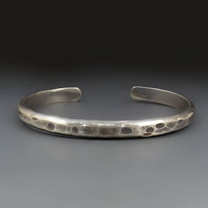 May include: A silver cuff bracelet with a hammered, textured finish and a polished sheen. The open-ended band features a series of small, rounded indentations along the outer surface, creating a tactile, irregular pattern across the metal.