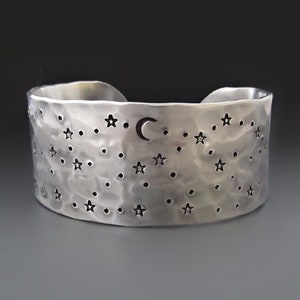May include: A silver cuff bracelet with a hammered texture. The bracelet features cut-out star and dot designs, with a crescent moon. The bracelet is approximately 2 inches wide.