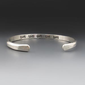 May include: A silver-colored metal cuff bracelet with a smooth, polished finish. The outer surface features five distinct black tally mark engravings spaced evenly along the band. The bracelet has an open design with rounded, tapered ends.