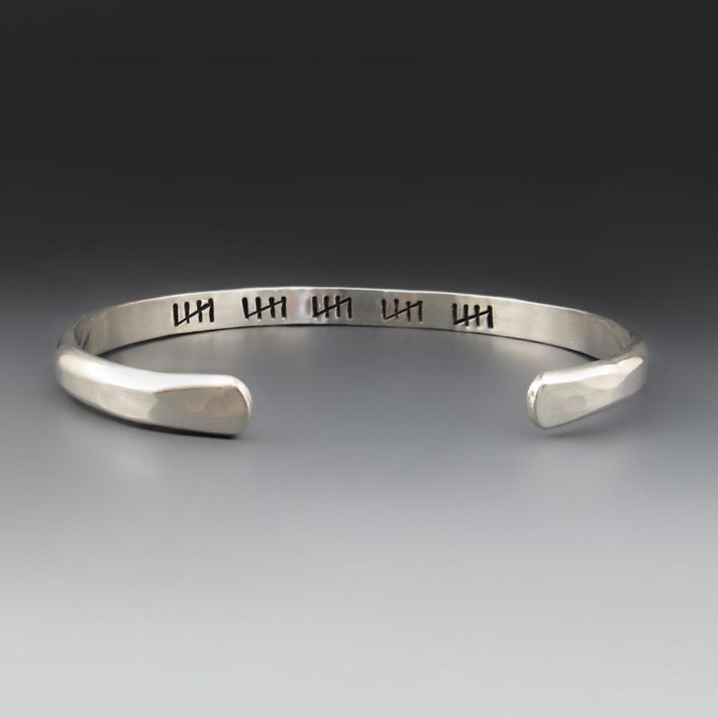 May include: A silver-colored metal cuff bracelet with a smooth, polished finish. The outer surface features five distinct black tally mark engravings spaced evenly along the band. The bracelet has an open design with rounded, tapered ends.