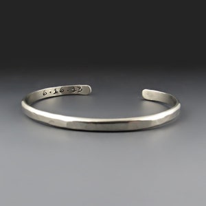 May include: A silver-toned metal cuff bracelet with a hammered texture. The interior surface of the bracelet features the stamped text 6.16.13 in a simple, dark font. The bracelet has an open design with rounded ends and a polished finish.