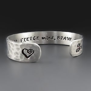 May include: A silver cuff bracelet with a hammered texture. The bracelet features a heart design and the engraved text: "FIERCE mind, BRAVE..." A Celtic symbol is on the opposite end. This jewelry item is a stylish accessory.