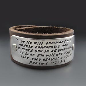May include: A dark brown leather cuff bracelet featuring a wide, curved silver-tone metal plate attached with two rivets. The plate is engraved with the text: "For He will command His angels concerning you to guard you in all your ways... so that you will not strike your foot against a stone." Psalms 91:11.