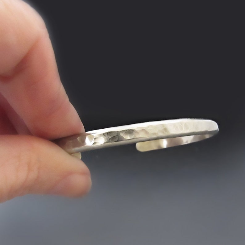 May include: A silver-colored metal cuff bracelet featuring a hammered, textured surface. The bracelet has an open design with a polished finish that reflects light across its uneven, dimpled exterior, creating a distinct visual pattern.