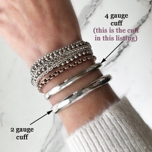 May include: A collection of silver-toned bracelets including three chain-link styles and two smooth metal cuffs. Text labels indicate a 4 gauge cuff and a 2 gauge cuff. The bracelets are stacked on a wrist, showcasing varying textures and thicknesses.