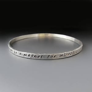 May include: A silver bangle bracelet with the inscription "A heart is a magnet for miracles".