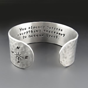 May include: Silver cuff bracelet with a hammered texture. The bracelet has an engraved compass design and the text "You already possess everything necessary to become great."