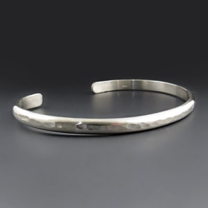 May include: A silver-colored metal cuff bracelet with a polished finish and a hammered texture along the outer surface. The bracelet features an open-ended design with rounded terminals, showcasing a reflective surface that catches the light.