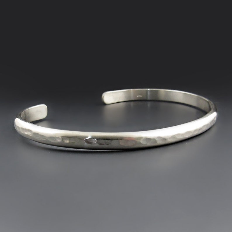 May include: A silver-colored metal cuff bracelet with a polished finish and a hammered texture along the outer surface. The bracelet features an open-ended design with rounded terminals, showcasing a reflective surface that catches the light.