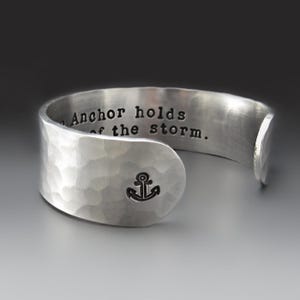 May include: A silver-toned cuff bracelet with a hammered texture. The exterior features a small stamped anchor icon. The interior is engraved with the text, The Anchor holds the storm.