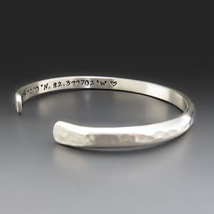 May include: A silver cuff bracelet with a hammered, textured exterior finish. The interior surface is engraved with the coordinates 34.2290 N, 82.399702 W followed by a small heart symbol. The bracelet has an open design for easy adjustment.