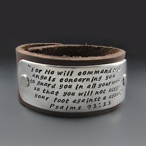May include: A brown leather cuff bracelet with a silver plate engraved with the text "For He will command His angels concerning you to guard you in all your ways... so that you will not strike your foot against a stone. Psalms 91:11"