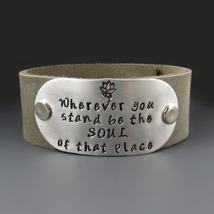 May include: A gray leather cuff bracelet with a silver oval charm that reads "Wherever you stand be the soul of that place". A small flower design is engraved above the text.