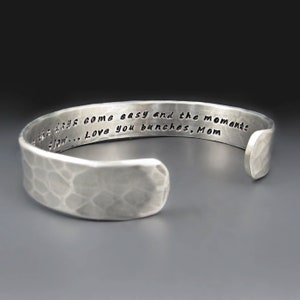 May include: Silver cuff bracelet with a hammered texture. The bracelet has an open design and features an engraved message: "...the days come easy and the moments flow... Love you bunches, Mom."