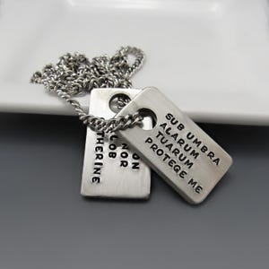 May include: A silver-toned metal pendant necklace featuring two rectangular dog tags suspended from a chain. The front tag is stamped with the text SUB UMBRA ALARUM TUARUM PROTEGE ME, while the partially visible rear tag displays the text ON NOR LOB THERINE.