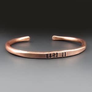May include: A copper bracelet with a hammered finish and a simple inscription of four vertical lines and two parallel lines.