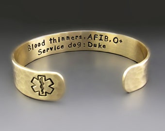 Puede incluir: Pulsera de alerta médica de oro con acabado martillado. La pulsera tiene el texto "Blood thinners, AFIB, O+ Service dog: Duke" grabado en el exterior y un símbolo médico en el interior.