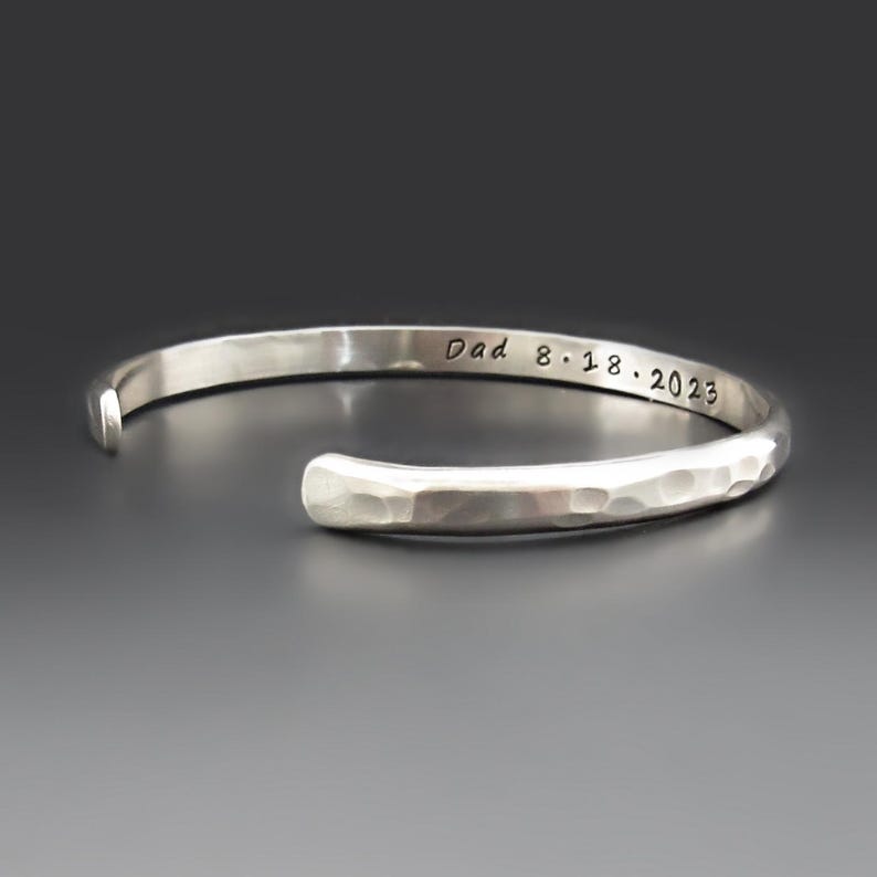May include: A silver-colored cuff bracelet with a hammered, textured finish on the exterior. The inner surface of the bracelet features the engraved text "Dad 8.18.2023" in a simple, dark, sans-serif font. The bracelet has an open design.
