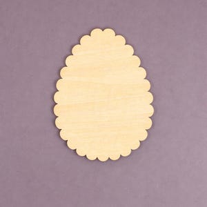 May include: A light-colored, wooden egg-shaped cutout with a scalloped edge. The cutout is made of unfinished wood and is set against a muted purple background. This craft supply is ideal for DIY projects and decorations.
