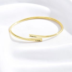 May include: A gold-colored, open cuff bracelet with the names "Dianna" and "Linda" engraved on the ends. The bracelet has a smooth, polished finish and is displayed on a white, textured surface.