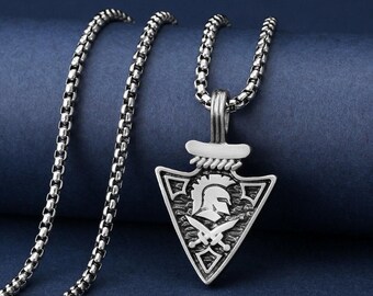May include: Silver-toned pendant necklace featuring a triangular pendant with a Spartan helmet design and crossed swords. The pendant hangs from a chain with a box link design. The necklace is displayed on a dark blue background.