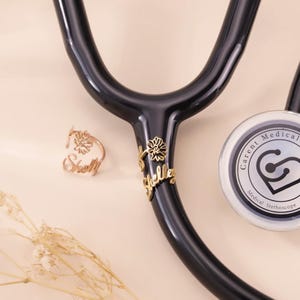 May include: A black stethoscope with a rose gold ring and charm. The ring is personalized with the name "Shelly" and a flower design. The charm on the stethoscope also has the name "Shelly" and a flower design. The stethoscope head has the text "Carent Medical".