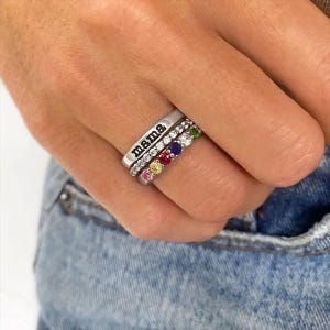 May include: Three silver rings stacked on a finger. The top ring has the word "mama" engraved on it. The middle ring is encrusted with clear stones. The bottom ring has a row of colourful gemstones: pink, yellow, red, purple, and green.