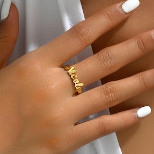 Può includere: Un anello color oro con il nome "Nicole" in corsivo. L'anello è indossato su un dito, con uno sfondo morbido e neutro. È un gioiello.