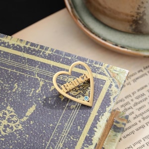 May include: A gold-coloured heart-shaped brooch with the name "Branca" inscribed across the centre. The brooch features a small, round, light blue gemstone and is resting on a book with a vintage design.