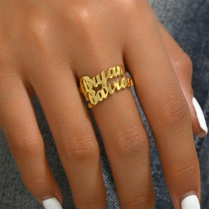 Puede incluir: Un anillo dorado con los nombres "Bryan" y "Barron" en fuente cursiva. El anillo se lleva en un dedo, mostrando el diseño personalizado. El anillo es una pieza de joyería.