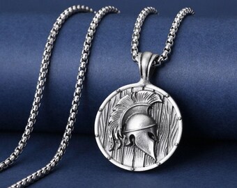 May include: Silver-toned pendant necklace featuring a detailed Spartan helmet design within a circular frame. The pendant hangs from a matching chain. The background is a solid, dark blue.