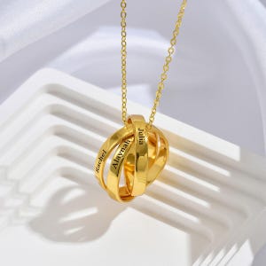 May include: A gold-colored necklace with three interlocking rings. Each ring is engraved with a name: Rachel, Alaynah, and Julia. The necklace hangs from a delicate chain, displayed on a white surface.