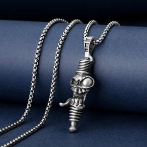 Könnte beinhalten: Silberfarbene Halskette mit Anhänger im stilisierten Schädel-Design. Der Anhänger hat ein detailliertes, strukturiertes Aussehen mit zylindrischem Sockel und einer Kette im Kettenglied-Stil. Der Hintergrund ist dunkelblau.