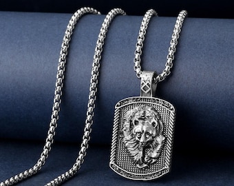 May include: Silver-coloured necklace featuring a rectangular pendant with a detailed lion's head design. The pendant is attached to a chain with a box link style. The necklace is displayed against a dark blue background.