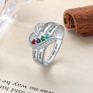 Puede incluir: Un anillo plateado con un diseño en forma de corazón, con cuatro piedras preciosas de colores y los nombres "Sammy", "Mamie", "Jackie" y "Kaley". El anillo está adornado con pequeñas piedras transparentes y la inscripción "Love each other".