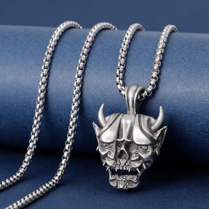 May include: A silver-colored pendant necklace featuring a detailed, horned demon skull design. The skull has sharp teeth and a grimacing expression. The chain is a linked style, and the background is a solid blue.
