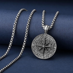 May include: Silver-colored compass pendant necklace. The pendant features a detailed compass rose design with directional markings. The chain is a matching silver color and has a textured, rounded link design. The necklace is displayed on a blue background.