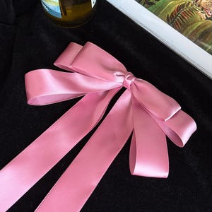 May include: A pink satin ribbon bow with long, flowing tails. The bow is tied in the center and lies on a black surface. The ribbon has a shiny, smooth texture and is a soft, pastel pink color.
