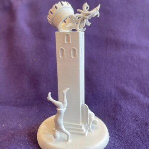 Può includere: Una scultura bianca stampata in 3D di una torre con una sommità decorativa e figure. Una figura fa una verticale e un'altra è seduta. La base della scultura è rotonda e riporta la scritta "TOWER".