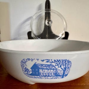 Puede incluir: Cuenco de cerámica blanco con una ilustración azul de una casa y un paisaje. Una tapa de cristal transparente está detrás del cuenco sobre un soporte negro. El cuenco está sobre una superficie de madera.