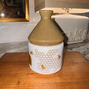 May include: A white and gold honey jar with a beehive-shaped lid. The jar features a honeycomb pattern with bees. The lid is a light brown color. The jar is sitting on a wooden surface.