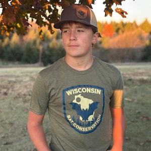 Puede incluir: Una camiseta verde oliva con un gráfico azul y blanco que dice "Wisconsin Bat Conservation". La camiseta se combina con una gorra de camionero marrón y beige con un parche de Wisconsin. La imagen está ambientada al aire libre con un fondo borroso.