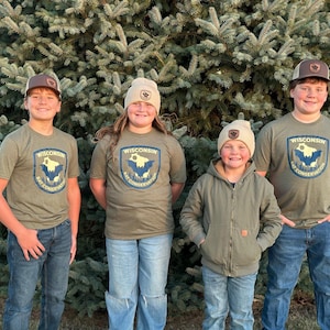 Op de afbeelding: Vier mensen dragen bijpassende olijfgroene t-shirts, hoeden en een jas. De shirts tonen een Wisconsin Bat Conservation-logo. De groep staat voor een grote groenblijvende struik. De hoeden zijn bruin en beige.