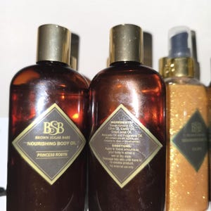 Puede incluir: Aceite corporal nutritivo Brown Sugar Babe en una botella de vidrio marrón con tapa dorada. La etiqueta tiene forma de diamante con el texto "BSB" y "Brown Sugar Babe - Nourishing Body Oil - Princess Robyn".