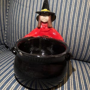 May include: A ceramic figurine of a witch wearing a black pointed hat with a yellow band and a red cloak, standing behind a glossy black cauldron. The witch has brown hair and a red collar, while the cauldron features two small side handles.