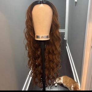 Puede incluir: Una larga peluca ondulada con raíces oscuras y reflejos castaños se exhibe en una cabeza de maniquí. La peluca está peinada con rizos sueltos y está montada en un soporte negro. El fondo incluye una pared gris y un vistazo a un pasillo.